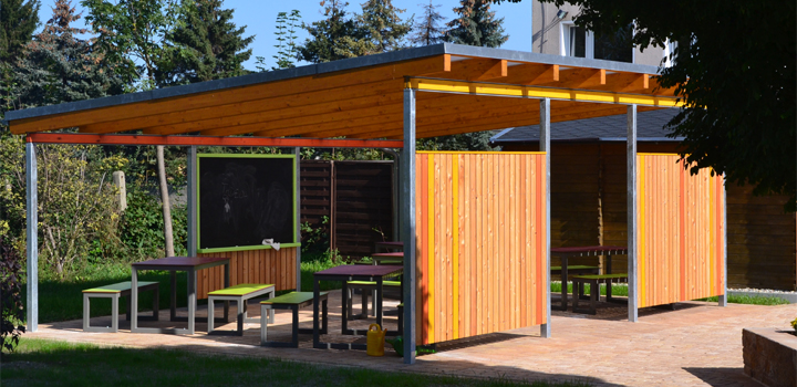 Wartehaus für eine Schule mit Holzwänden, Metallrahmen, Tafel, Tisch und bunten Bänken, von FreiraumObjekte Ney GmbH.