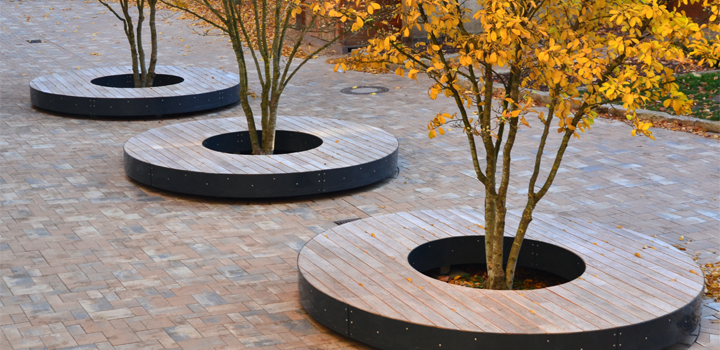 Mehrere runde Holzbänke mit Metalleinfassung um Bäume mit herbstlichem Laub, möglicherweise von FreiraumObjekte Ney GmbH.